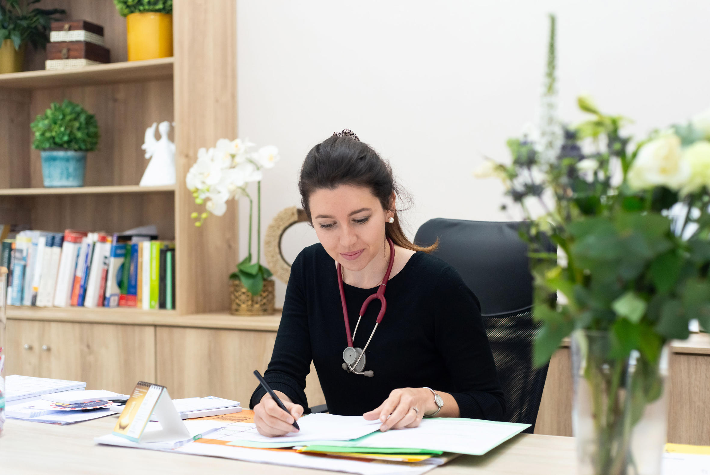 Female doctor with a stethoscope around her neck writing on documents at a desk in an office with bookshelves and plants.