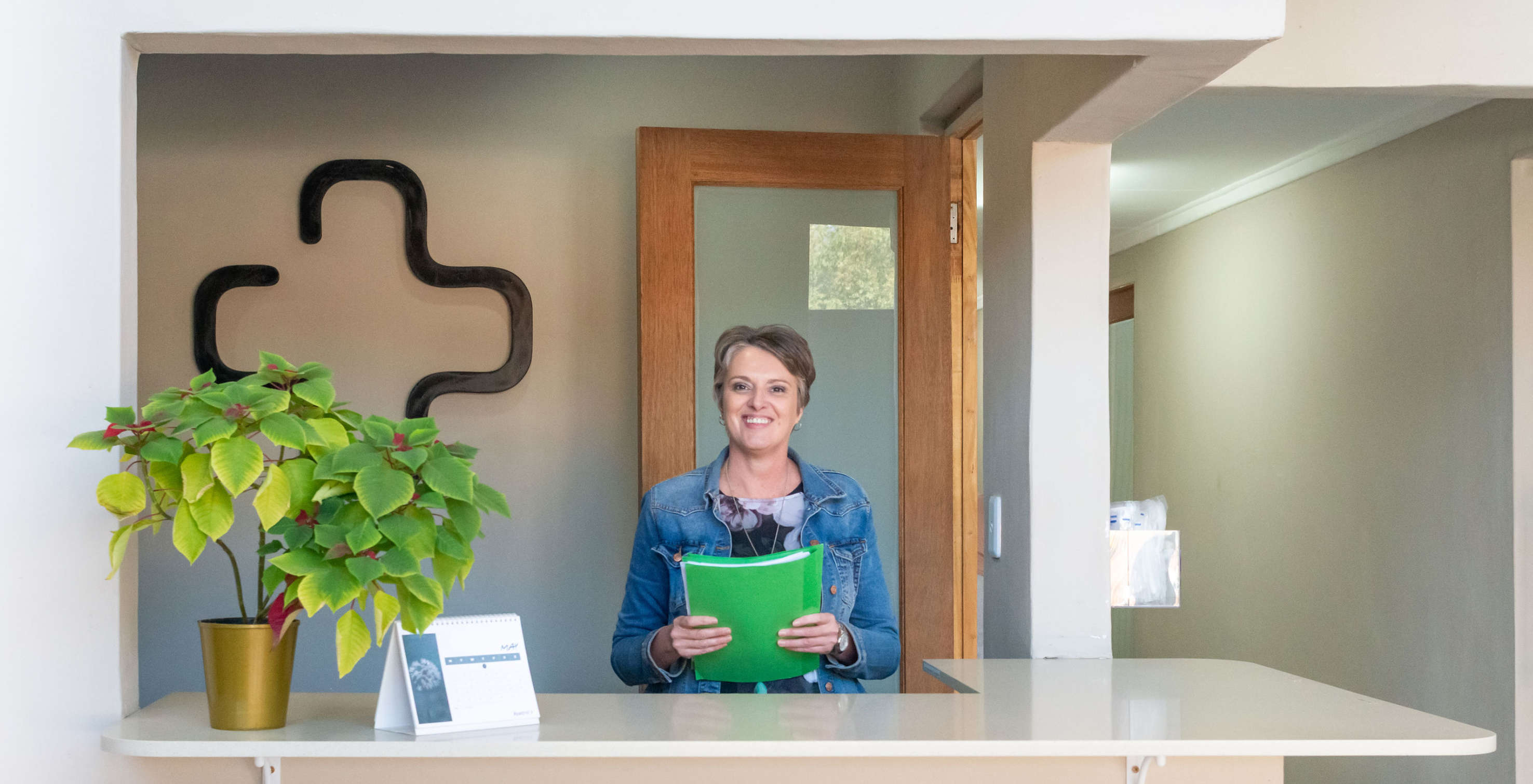 Woman smiling behind a reception desk holding a green folder, with a calendar and a potted plant on the counter, and a black cross decoration on the wall behind her.