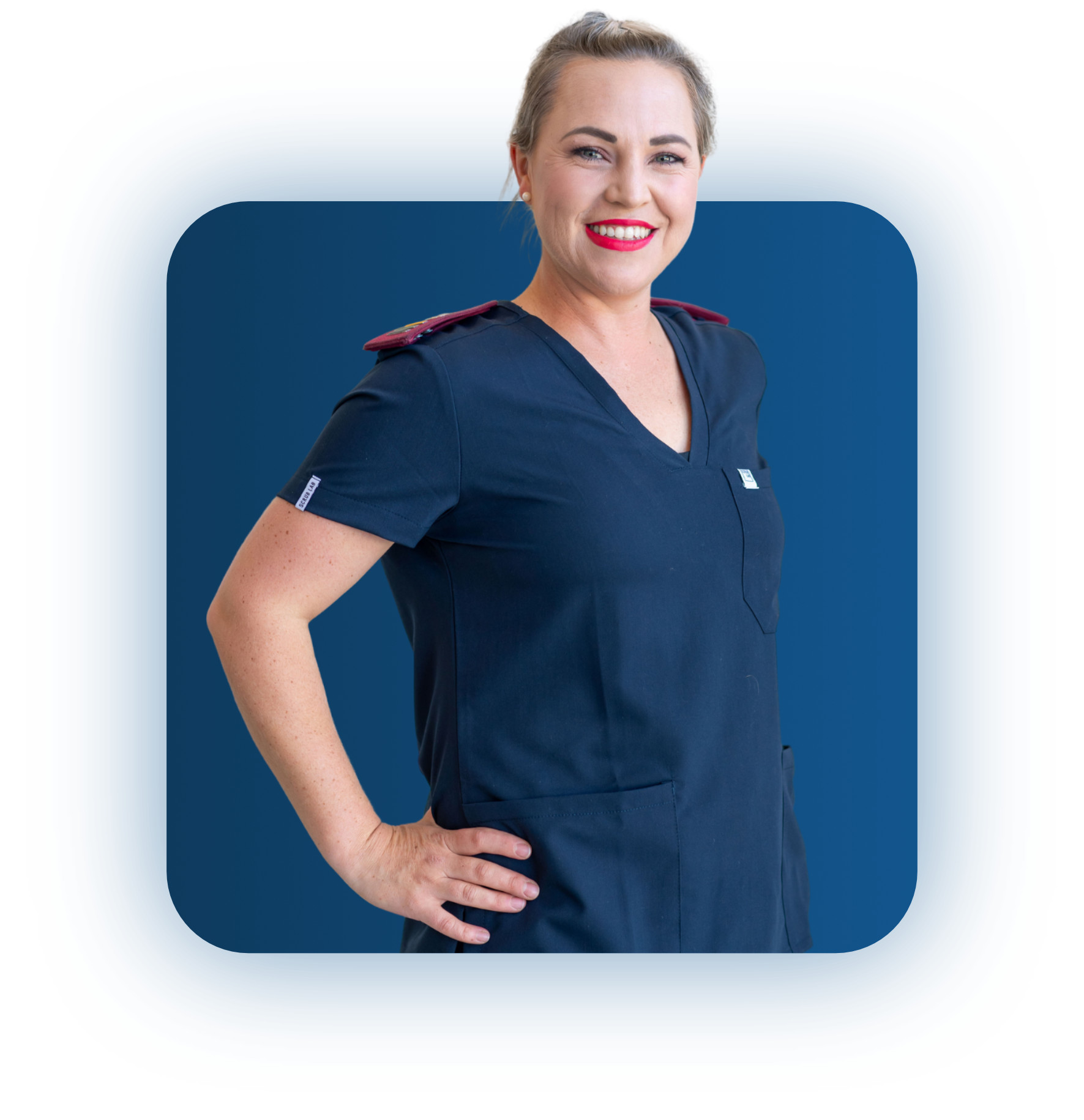 Smiling woman with light hair in a navy blue medical scrub top with red shoulder epaulets, standing with one hand on her hip against a blue background.