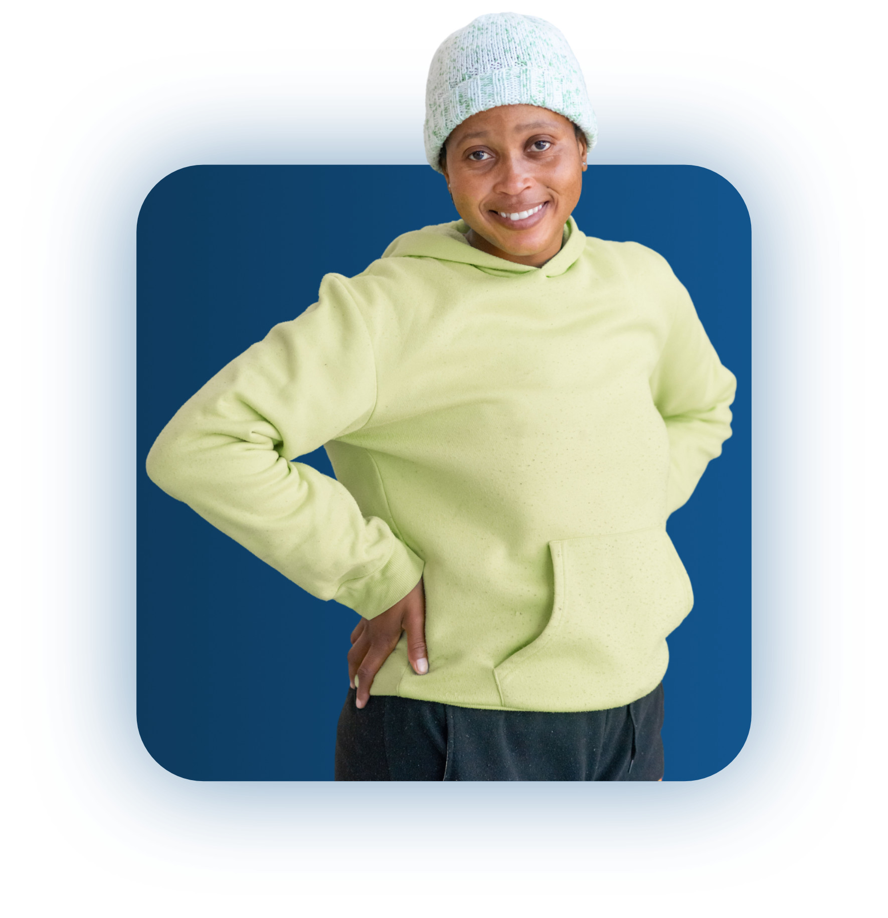 Smiling person wearing a light green hoodie and knitted beanie, standing with hands on hips against a blue background.